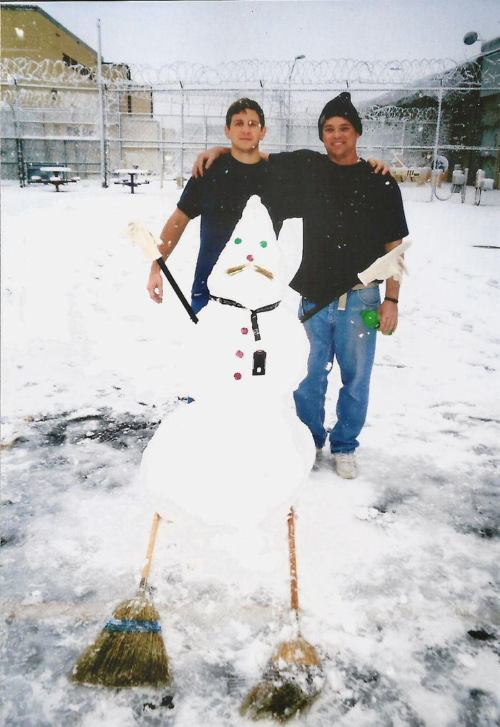 Two men standing either side of a snowman built in a snow-covered prison yard, razor wire fencing behind them